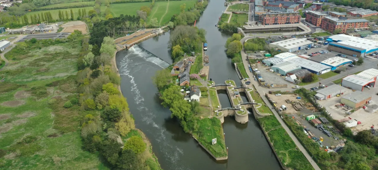 Aerial view of the River Severn winding through Worcester past the weir and locks at Diglis island, with green riverside meadows on one bank and the city and cathedral beyond.